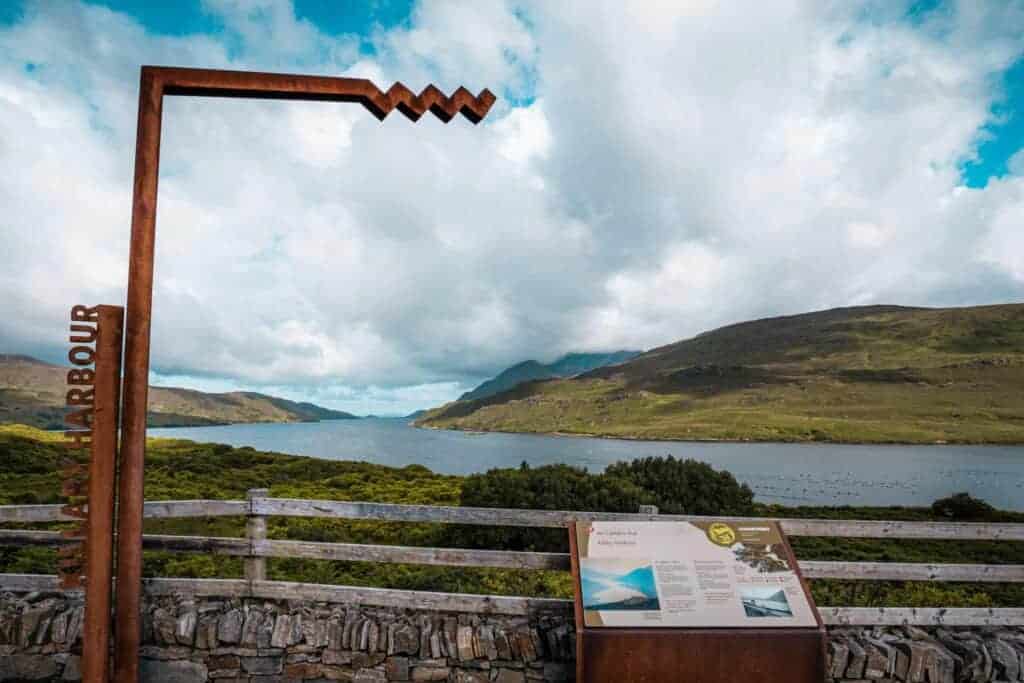 iew of Killary Harbour framed by a rusted metal structure and an information sign, with lush green hills and a partly cloudy sky in the background