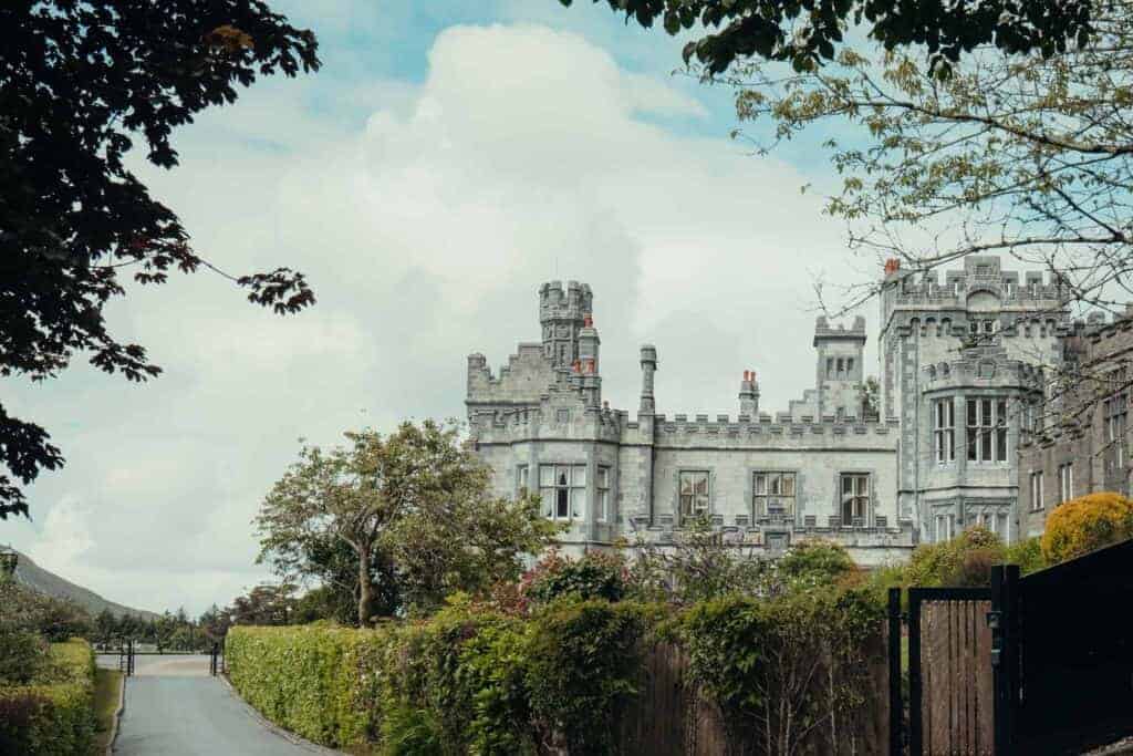 A wide view of Kylemore Abbey against a backdrop of green hills and trees, with a lake in the foreground.