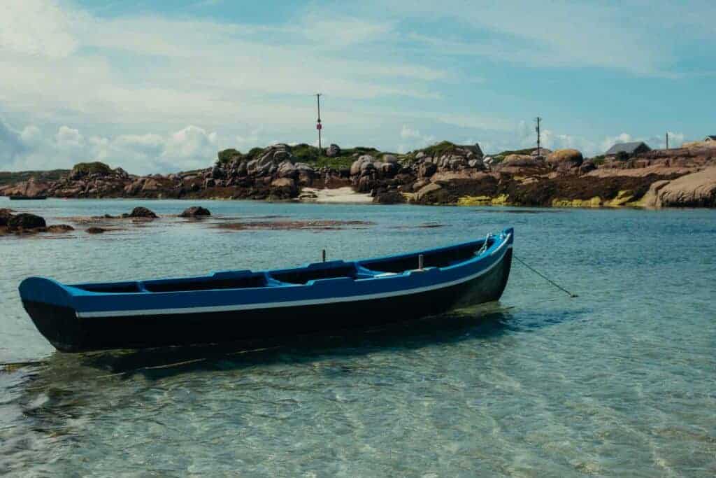 A blue boat anchored in clear, shallow water near a rocky shore and small houses in the background.