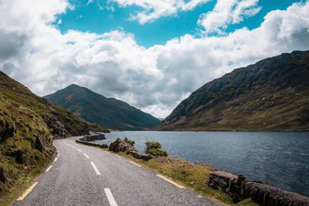A winding road runs alongside Doolough Lake, with mountains and a partly cloudy sky creating a dramatic landscape.