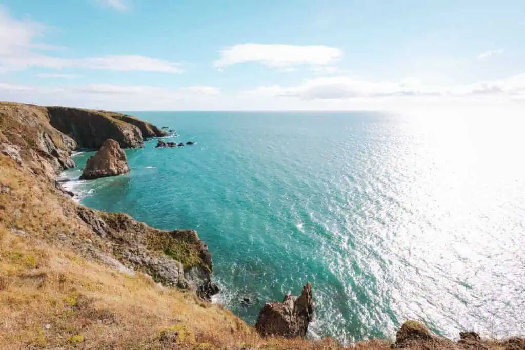 A stunning view of Ireland's Copper Coast, showcasing rugged cliffs and rocky outcrops that extend into the vibrant turquoise ocean. The sun shines brightly, casting a shimmering reflection on the water, while grassy cliffs slope down toward the sea. The coastline stretches into the distance under a blue sky with scattered clouds.