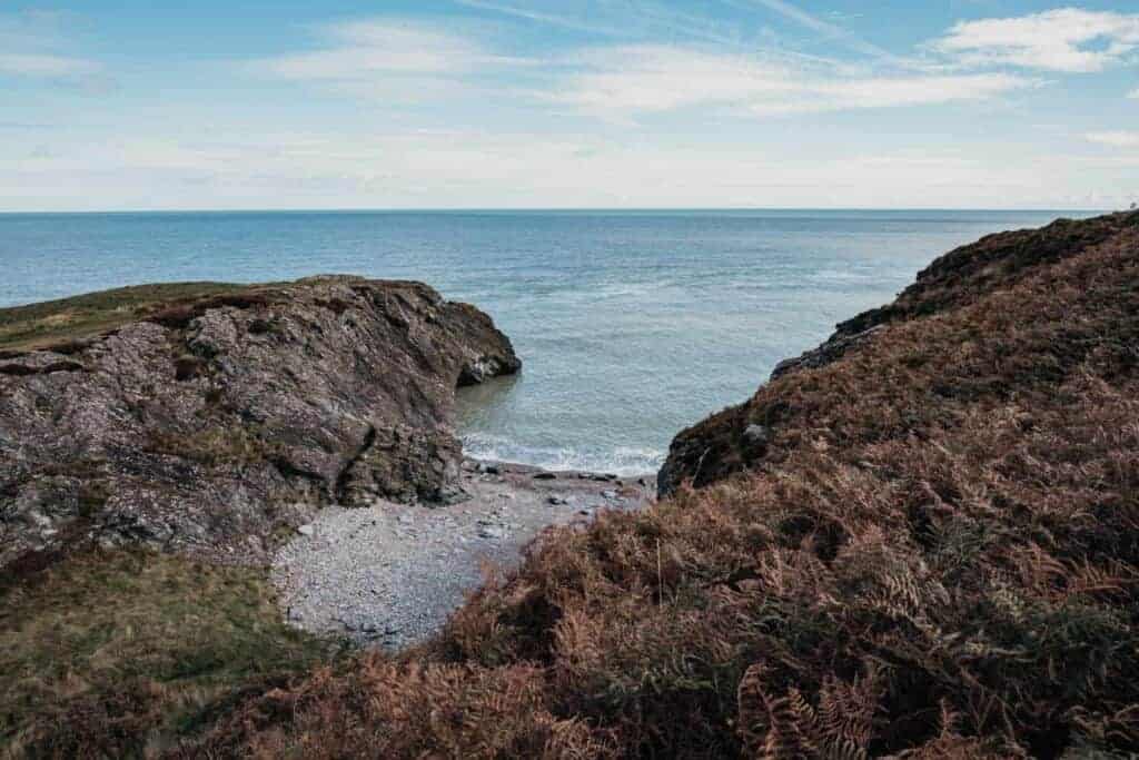 A small, pebbly cove nestled between rocky cliffs with calm waters stretching out to the horizon under a partly cloudy sky.