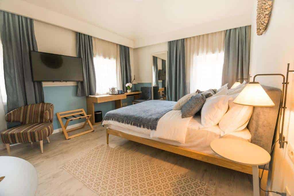 Cozy boutique hotel room close to Budva Old Town featuring a neatly made bed with layered pillows, soft lighting, and large curtained windows. A desk, wall mounted TV, and modern furnishings create a comfortable stay near the Adriatic coast.