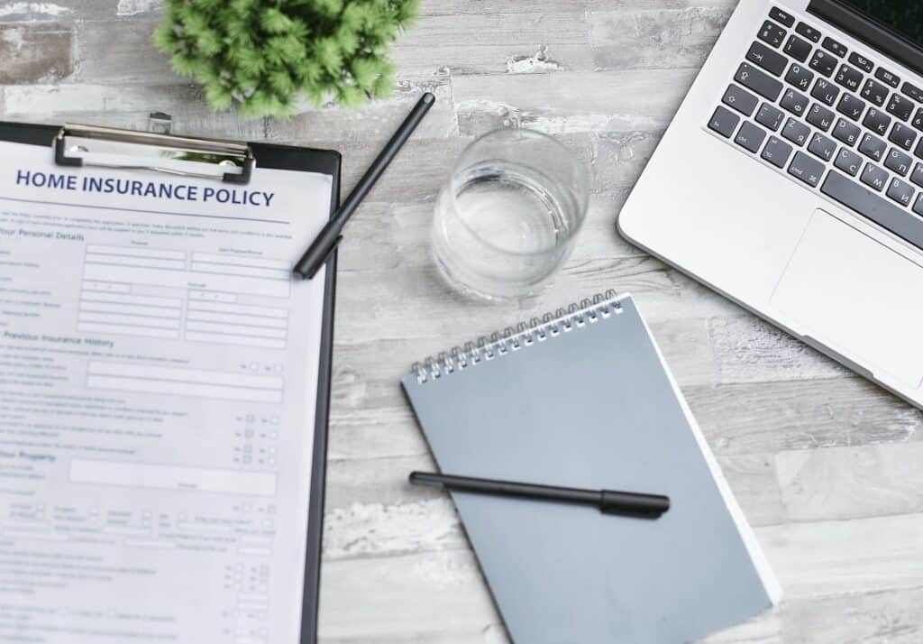Top-down view of a light wooden desk with a home insurance policy clipboard, a laptop, a notebook, two black pens, a glass of water, and a small green plant