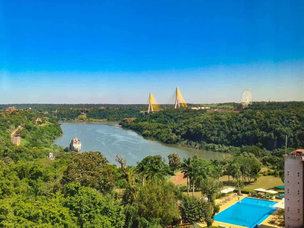 The photograph displays a scenic view from the hotel room in Puerto Iguazu, showcasing a serene river flanked by lush greenery. A prominent bridge with distinctive yellow triangular structures spans the river in the distance. On the right, a large Ferris wheel creates a focal point against the clear sky. In the foreground, a swimming pool with azure water provides a leisurely contrast to the natural scene.