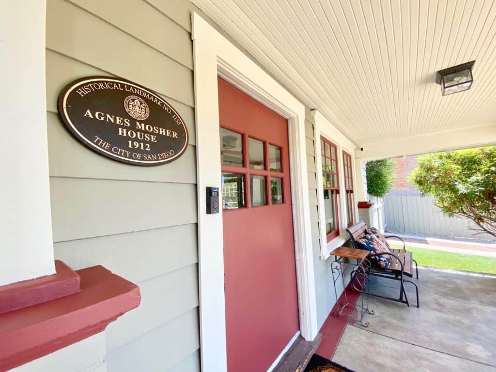 Close-up of a historical landmark plaque for the Agnes Mosher House (1912) mounted on grey horizontal siding next to a red craftsman front door