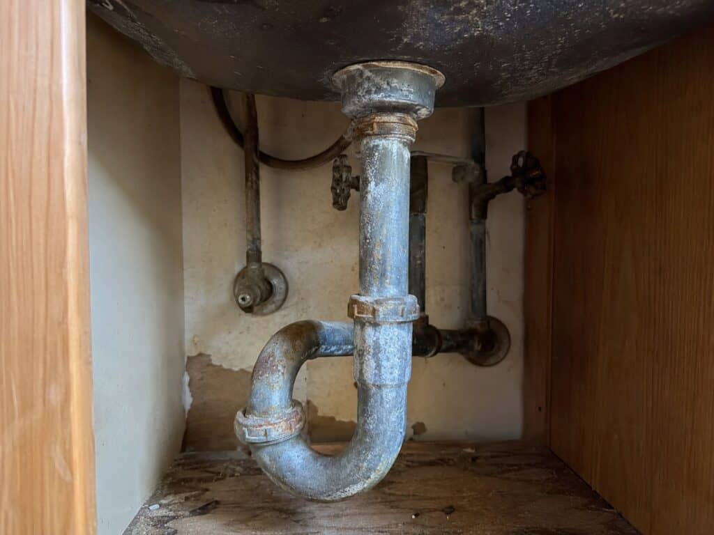 Corroded galvanized steel plumbing pipes under a kitchen sink in an older 1920s bungalow.