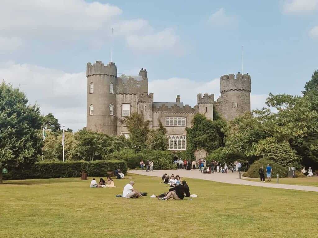 A view of Malahide Castle in Ireland, showcasing its grand stone structure with rounded towers and ivy-covered walls. The castle is set against a backdrop of a clear blue sky, with visitors relaxing and picnicking on the expansive green lawn in front of the entrance.