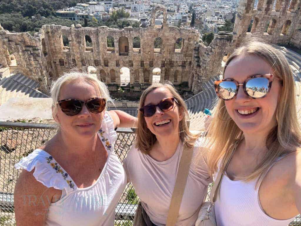 Three smiling women pose in front of the ancient stone ruins of an amphitheater, likely in Europe, enjoying a sunny sightseeing day. A joyful moment highlighting the social perks of travelling solo as a woman and connecting with fellow explorers.