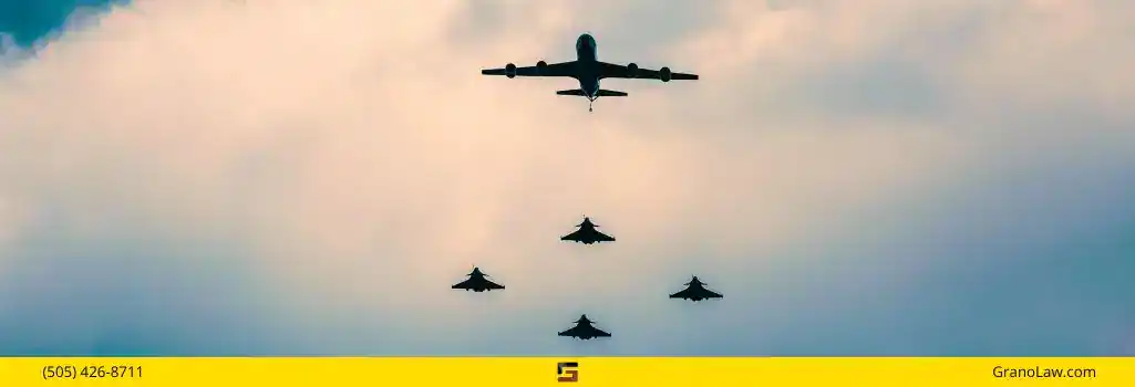A formation of five military aircraft flying in the sky. A large tanker aircraft is at the top, leading four fighter jets below it. The sky is cloudy and overcast. The image footer has a yellow bar with "(505) 426-8711" on the left, a logo in the center, and "GranoLaw.com" on the right.
