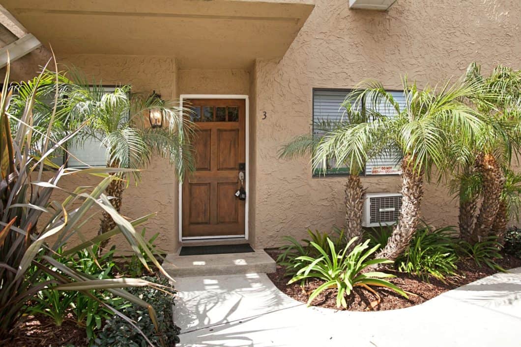 Condo entrance at 10154 Camino Ruiz featuring a classic wood paneled front door with a glass-paneled top, set within a tan stucco exterior and surrounded by tropical landscaping with small palm trees and lush greenery.