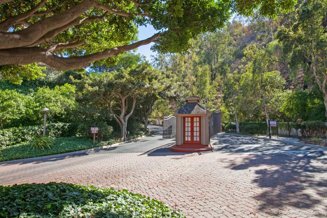 Gated community entrance at 5474 Caminito Borde featuring a brick-paved driveway, a security kiosk with red doors, and lush green hillside landscaping.