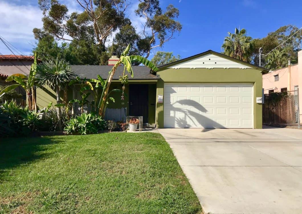 Vibrant olive-green duplex at 1908-10 31st Street in South Park featuring a two-car garage, lush tropical landscaping with banana trees, and a wide concrete driveway under a clear blue sky.