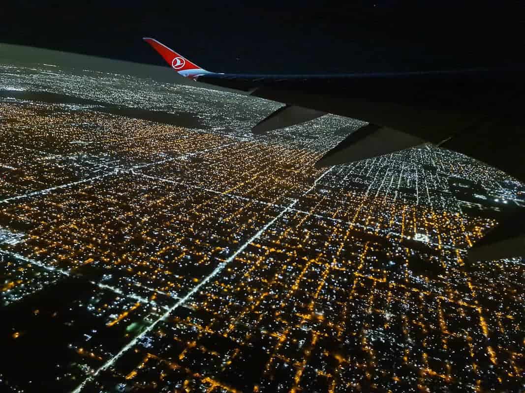 A night-time aerial view of a sprawling cityscape illuminated by countless lights, as seen from an airplane with the wingtip featuring a red logo visible against the dark sky.