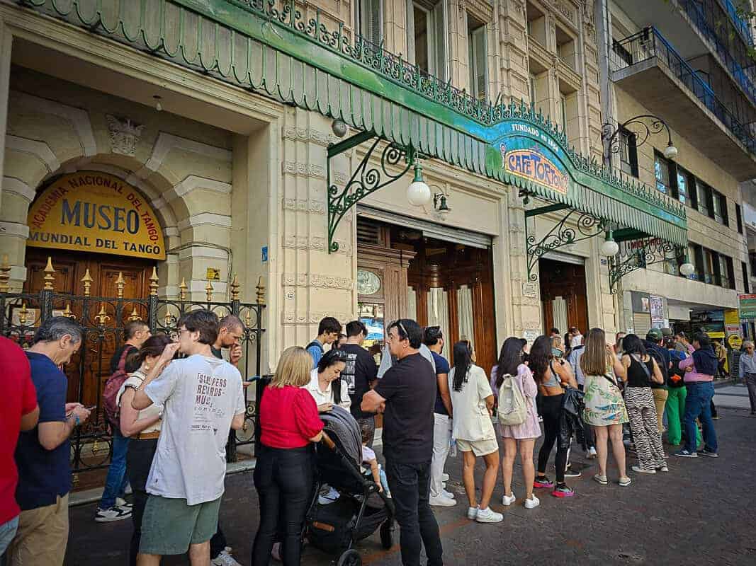 The image shows a bustling scene in front of a building with significant cultural heritage. There's a crowd of people who are waiting in line. The entrance to the building is adorned with an arch and decorative iron gates, and above, signs indicate the presence of both the "Academia Nacional del Tango" and the "Museo Mundial del Tango." Adjacent to this is a sign for the "Café Tortoni," a historically significant café known for its association with the tango. The café's façade features a traditional green awning. It seems to be a sunny day, and the crowd includes people of various ages, some looking at their phones, others engaged in conversation and all of them waiting to enter Cafe Tortoni.