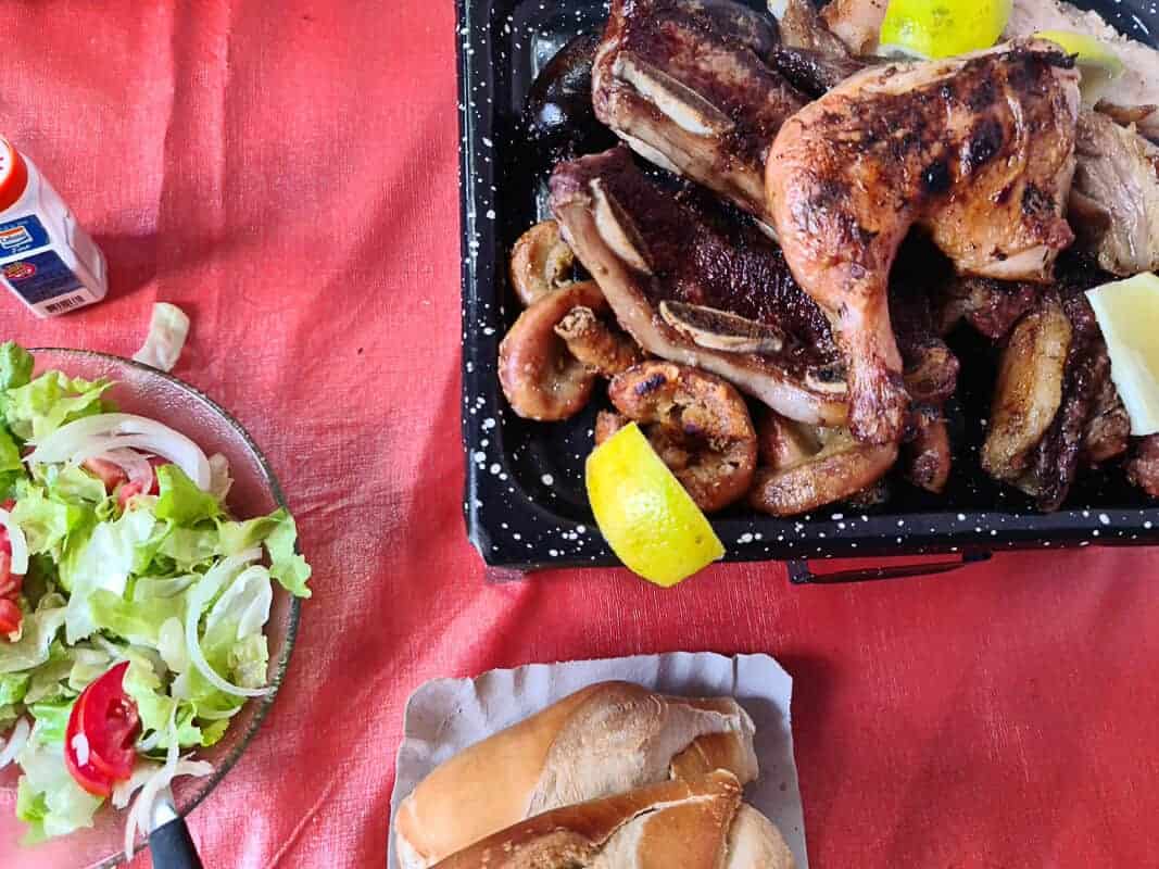 An appetizing Argentinian barbecue meal spread out on a table, featuring grilled meats such as steak and sausage, complemented by lemon wedges for added flavor. Accompanying the hearty proteins are a fresh salad with lettuce and tomato, along with a selection of bread rolls. This setup is typical of a casual outdoor dining experience, showcasing the rich culinary tradition of Argentina.