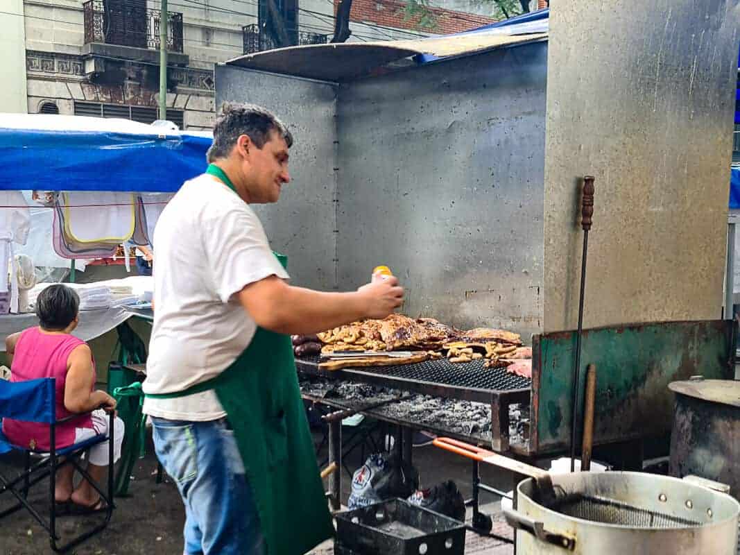 A man in a green apron is grilling a variety of meats at a busy street food stall af Feria da Mataderos Buenos Aires, spraying seasoning on them. The grill is full, suggesting a popular spot, possibly at an outdoor market or fair. Behind him, there are people seated under a blue tarp, indicating a casual dining environment common in outdoor markets. Smoke and the presence of a large pot, perhaps with oil for frying, contribute to a lively culinary scene.