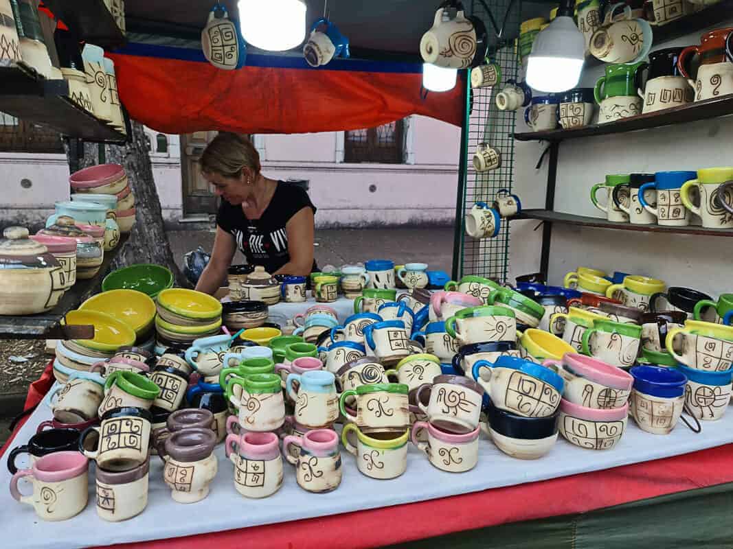 A vendor at Mataderos market stall arranges a colorful collection of handmade pottery. The ceramics feature playful designs and come in a variety of shapes and sizes, including mugs, bowls, and pots, with a mix of vibrant and pastel colors. The display is organized to showcase the unique craftsmanship of each piece, inviting passersby to take a closer look and perhaps choose a handmade item to take home.