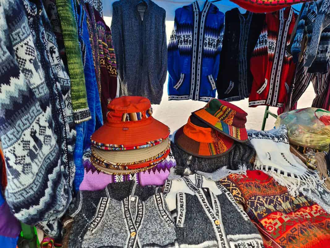 A market stall richly adorned with traditional handcrafted textiles. Woolen ponchos with geometric patterns hang in the background, exhibiting a variety of colors from deep blues to vibrant reds. In the forefront, artisanal hats with intricate woven bands rest atop a stack of more textiles, highlighting the detailed craftsmanship. The display offers a glimpse into the cultural fashion and skilled workmanship of the local artisans.
