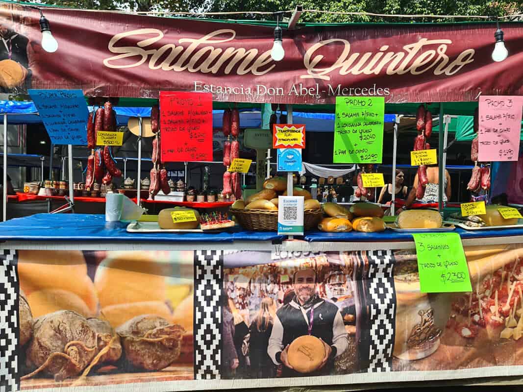 A colorful food stall at Feria de Mataderos named "Salame Quintero" from Estancia Don Abel - Mercedes. The stall features an array of homemade food products, prominently displaying various types of salami and cheese, with prices and promotions indicated on bright signs. Behind the counter, the reflection of the vendors can be seen in a mirror, and the backdrop features imagery of salami, contributing to the inviting presentation of the stall. This setup is typical of a traditional market, where local vendors sell their artisanal produce.