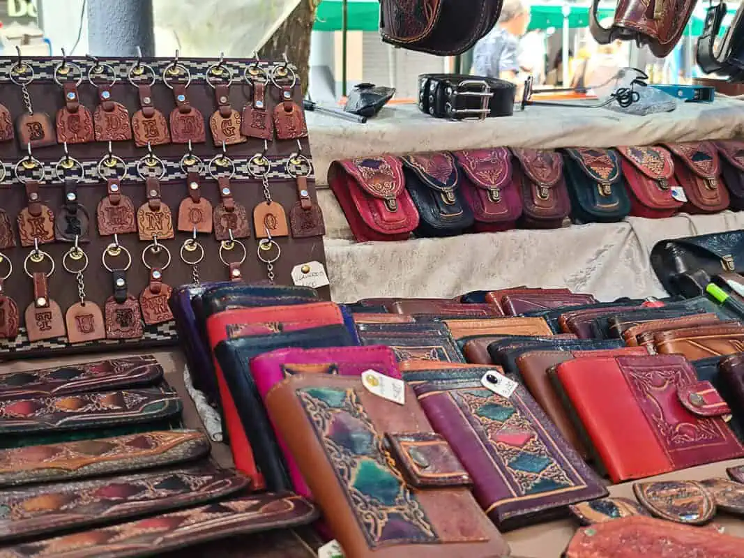 At a market stall, there's an assortment of handcrafted leather goods. The display features intricately embossed leather wallets in various hues like browns, reds, and blues, alongside small, ornate purses. Above the wallets, there are keychains with leather tags, each embossed with a different letter of the alphabet, hanging neatly in rows. This showcase of leather craftsmanship highlights the quality and cultural artisanship available at this market.