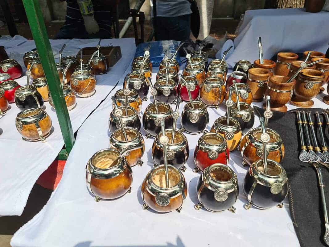A market stall displays a variety of mate cups, or "mates," traditionally used for drinking the South American herbal tea, mate. The collection showcases cups with various designs, including those with metal rims and decorative detailing. The mate cups are accompanied by bombillas, the special straws used for drinking mate. The arrangement also includes some wooden bowls and utensils, highlighting the rich cultural tradition of mate drinking in regions like Argentina.
