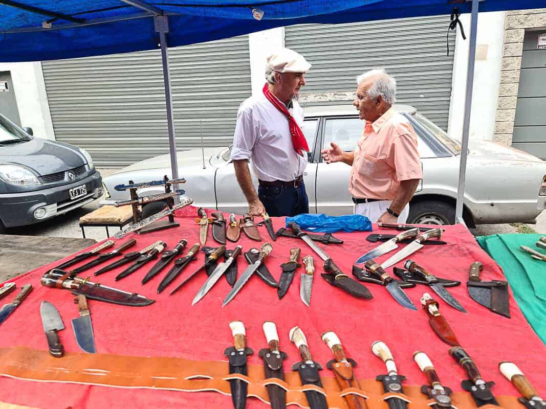 Two men engage in a conversation at a market stall that features a collection of traditional knives with various handle designs, laid out on a red cloth. The knives come in different sizes and appear to be handcrafted, with some displayed in leather sheaths. This kind of stall is characteristic of markets that sell handmade tools and crafts, reflecting local customs and craftsmanship.