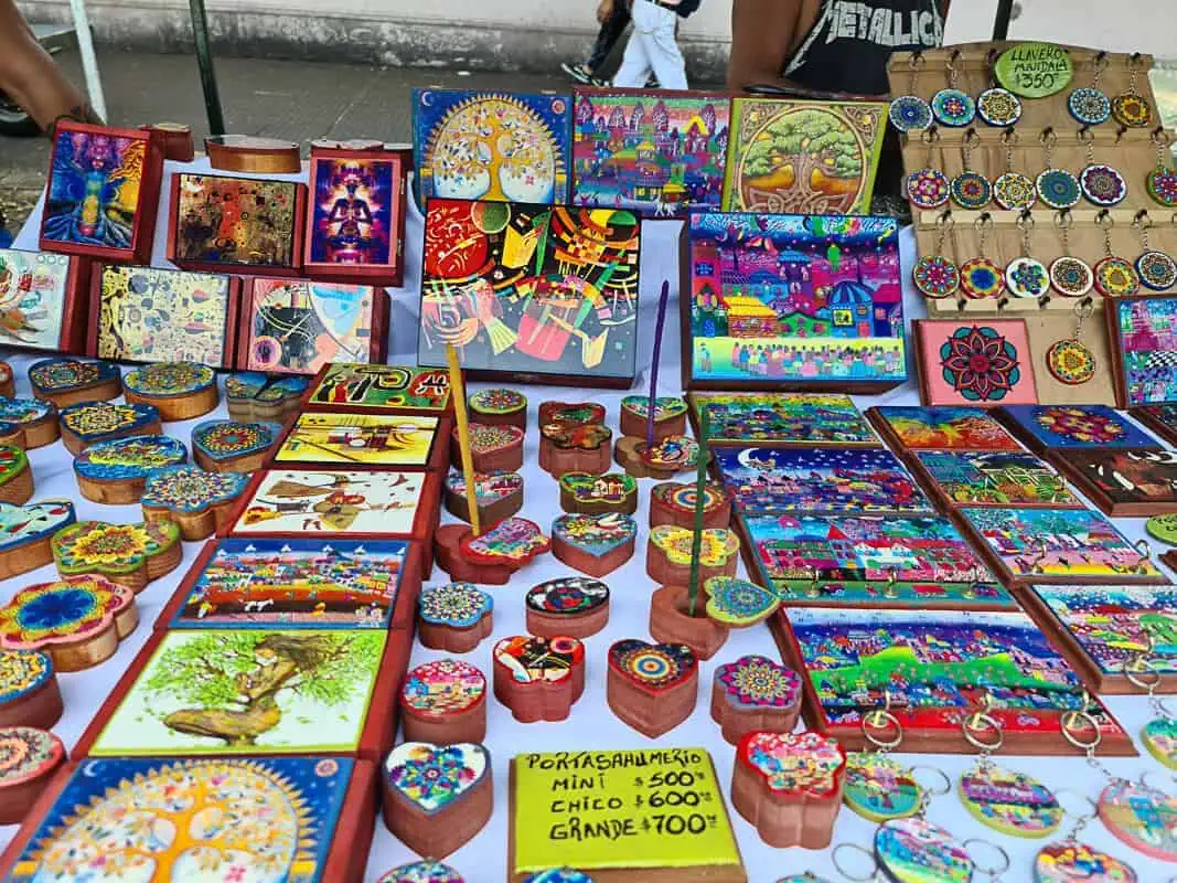 A vibrant array of handcrafted items is displayed on a table at Mataderos Fair, featuring intricate designs and bright colors. The collection includes various painted wooden boxes, possibly for jewelry or keepsakes, alongside decorative ceramic pieces such as coasters and trinkets. Artworks depicting colorful scenes and mandala designs are also visible, along with keychains that showcase similar patterns. Price tags suggest this is a market setup, inviting customers to purchase these unique, artisanal creations.