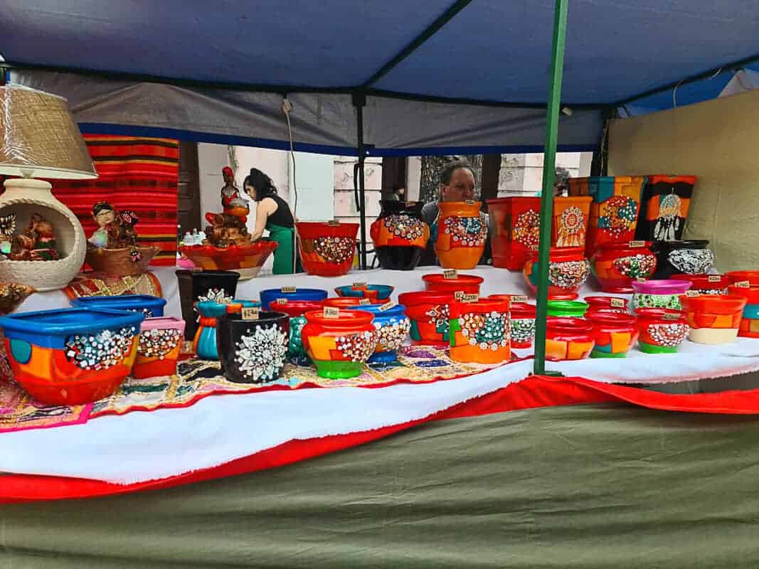Feria de Mataderos stall is richly adorned with brightly colored pottery. These hand-painted ceramic pots and bowls feature vibrant orange and blue hues, accented with intricate white floral and traditional patterns. The pieces vary in size and are thoughtfully arranged to create a visually striking display, inviting market-goers to browse and purchase. A vendor can be seen in the background, tending to the stall and the handcrafted wares.