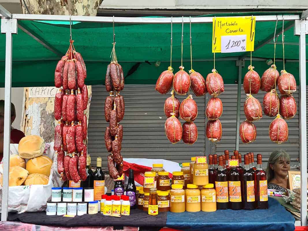 A market stall showcases an assortment of cured meats, including salami and mortadella, hanging in neat rows. Below, the table is lined with various bottles and jars, including honey and possibly homemade sauces or syrups. A sign highlights "Mortadela Caserita," indicating homemade quality, with a price tag for shoppers. Fresh loaves of bread also feature in the display, complementing the meats. The vendor, partially visible, adds to the lively market scene, offering a slice of local culinary tradition.