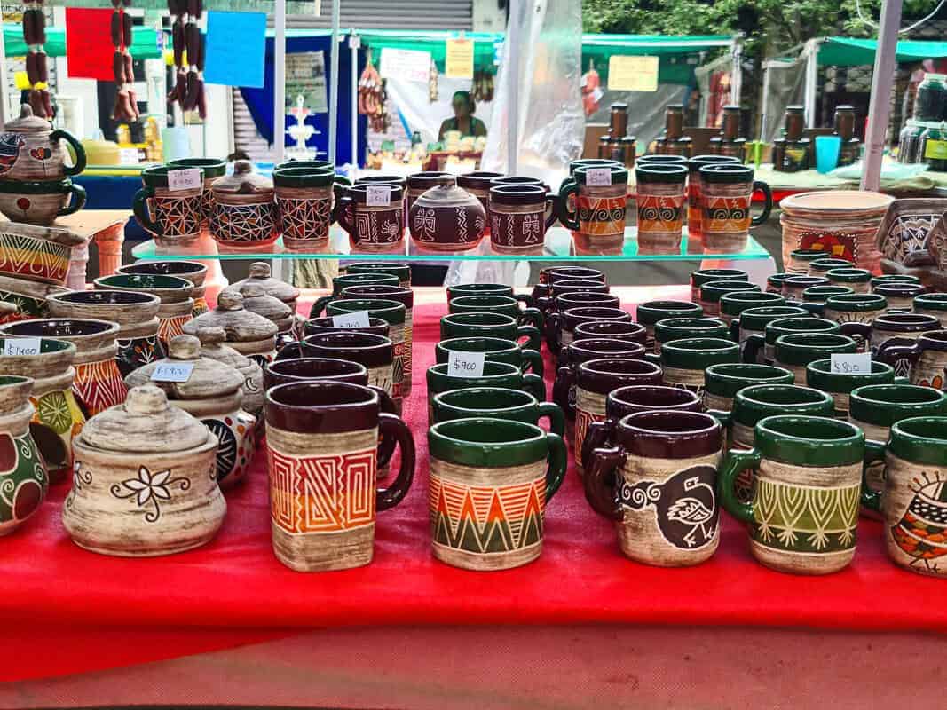 A collection of handmade ceramic mugs is on display at a market stall. Each mug features earthy tones and traditional patterns, possibly inspired by indigenous or folk designs. Some of the mugs have lids, and they vary in color, with some exhibiting natural clay hues and others in deep greens and browns. Price tags attached to each item suggest that these mugs are not only decorative but also functional and for sale, inviting market-goers to purchase a piece of local craftsmanship.