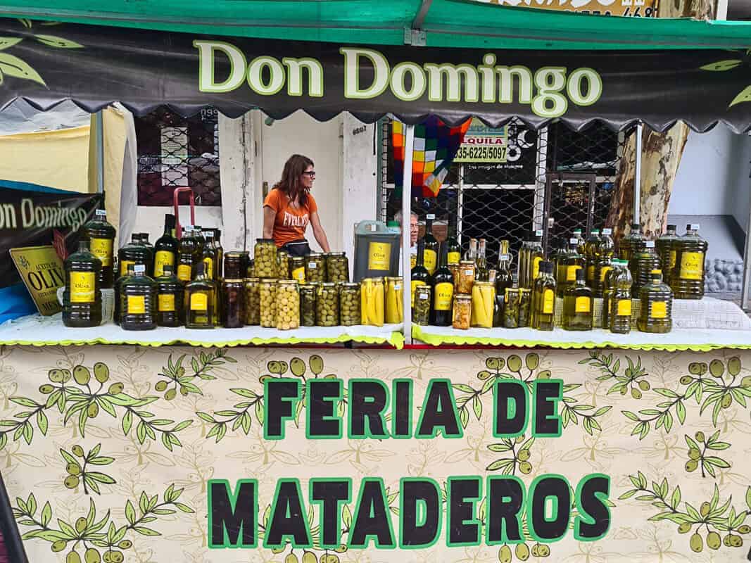 A vendor stands behind a stall at the Feria de Mataderos, displaying a selection of olive oils and olives. The signage "Don Domingo" indicates the brand or name of the stall, and the products range from bottles of olive oil to jars of olives, some possibly marinated or flavored. The olive-themed tablecloth adds to the presentation, emphasizing the natural, homemade quality of the products. The text "FERIA DE MATADEROS" is prominently displayed, anchoring the scene in this well-known market celebrated for its local produce and goods.
