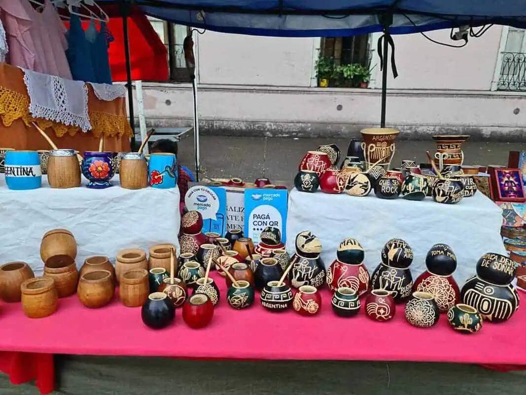 A market stall at Mataderos Fair displays an assortment of traditional Argentinian mate utensils. Various calabash gourds and wooden mate cups, some ornately decorated with colorful patterns and national symbols, are lined up alongside bombillas (metal straws), each designed for sipping mate, a cultural beverage. Additional crafts and decorative items featuring Argentinian motifs suggest a vibrant celebration of local traditions and craftsmanship at the fair.