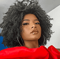 Uma mulher negra com cabelo crespo volumoso preto usa um top vermelho vibrante, brincos de argola grandes e maquiagem, olhando diretamente para a câmera com confiança.