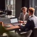 Dois homens de negócios conversam em uma mesa de madeira com um laptop aberto, um deles apontando para a tela, em um escritório moderno e bem iluminado.