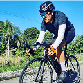 Um ciclista masculino com capacete preto, óculos de sol e uniforme azul pedala uma bicicleta preta em uma estrada ensolarada com árvores verdes e céu azul.