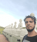 Um homem barbudo tira uma selfie à direita, com uma longa fileira de estátuas brancas de Buda sentadas ao ar livre sob um céu claro.