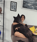 Uma mulher sorridente abraça uma grande criatura peluda escura, parecida com um lobo, sentada em um sofá cinza em uma sala de estar.