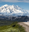 Uma majestosa montanha coberta de neve domina o horizonte, com picos rochosos e um vale verdejante atravessado por uma estrada de terra em primeiro plano.