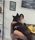 Uma mulher sorridente abraça uma grande criatura peluda escura, parecida com um lobo, sentada em um sofá cinza em uma sala de estar.