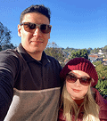Casal sorridente de óculos escuros e roupas de inverno tira uma selfie ao ar livre, com céu azul e paisagem urbana ao fundo.