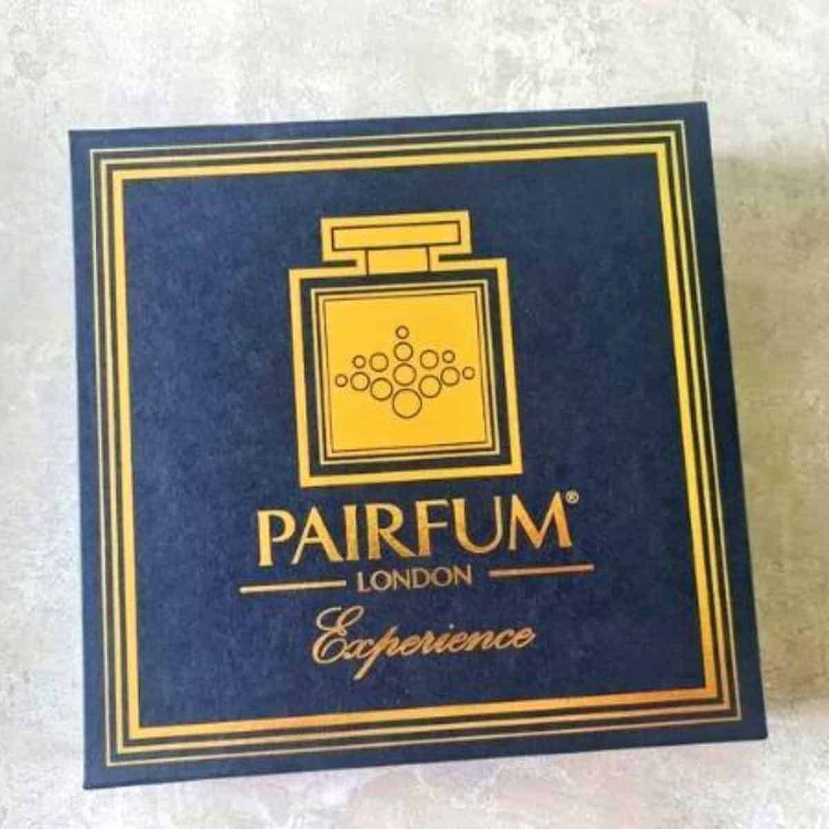 A box of Pairfum London Experience perfume rests elegantly on a light surface, showcasing its sophisticated gold and dark blue design.