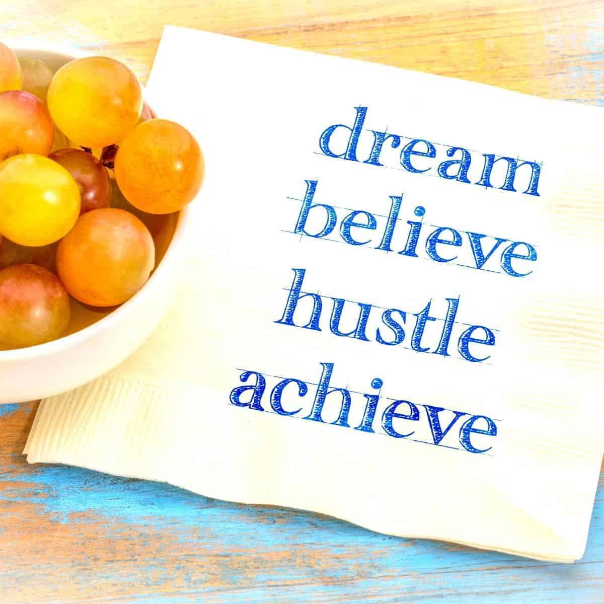 A bowl of grapes sits next to a napkin emblazoned with the mantra "dream, believe, hustle, achieve," embodying the essence of hustle culture.