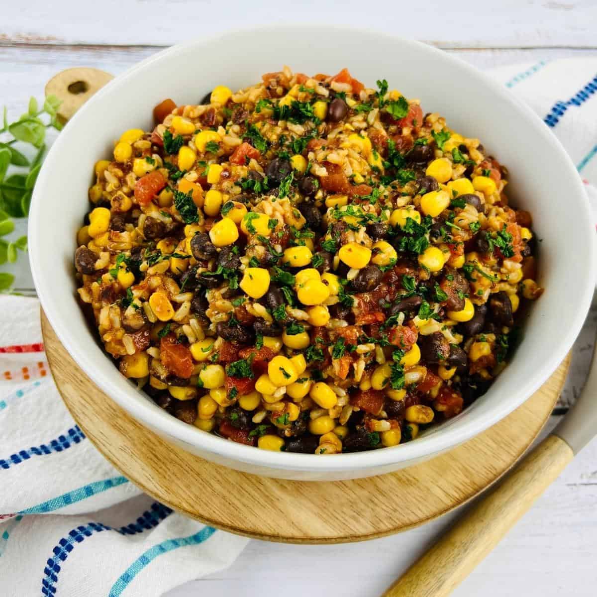 A bowl of vibrant tex mex salad with mixed corn, black beans, tomatoes, and herbs sits on a wooden board. A patterned napkin and spoon lie beside it, inviting a delicious dive into this quinoa-infused delight.