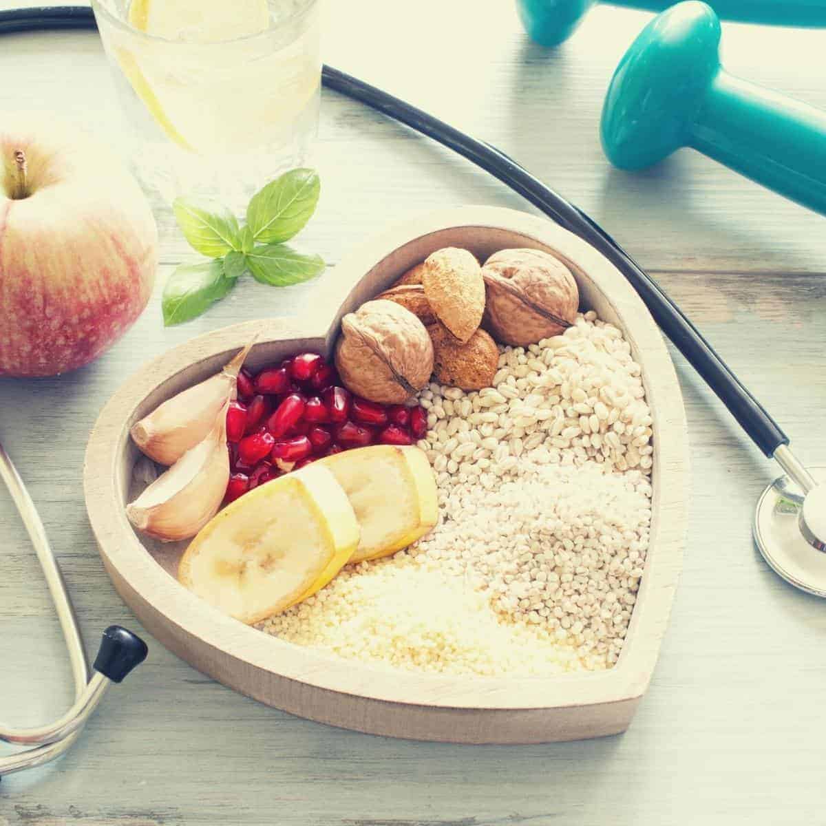 A heart-shaped bowl features foods to lower cholesterol, like nuts, pomegranate seeds, and grains. Scattered around are garlic, banana slices, an apple, mint leaves, water with lemon, hand weights, and a stethoscope on a wooden surface.