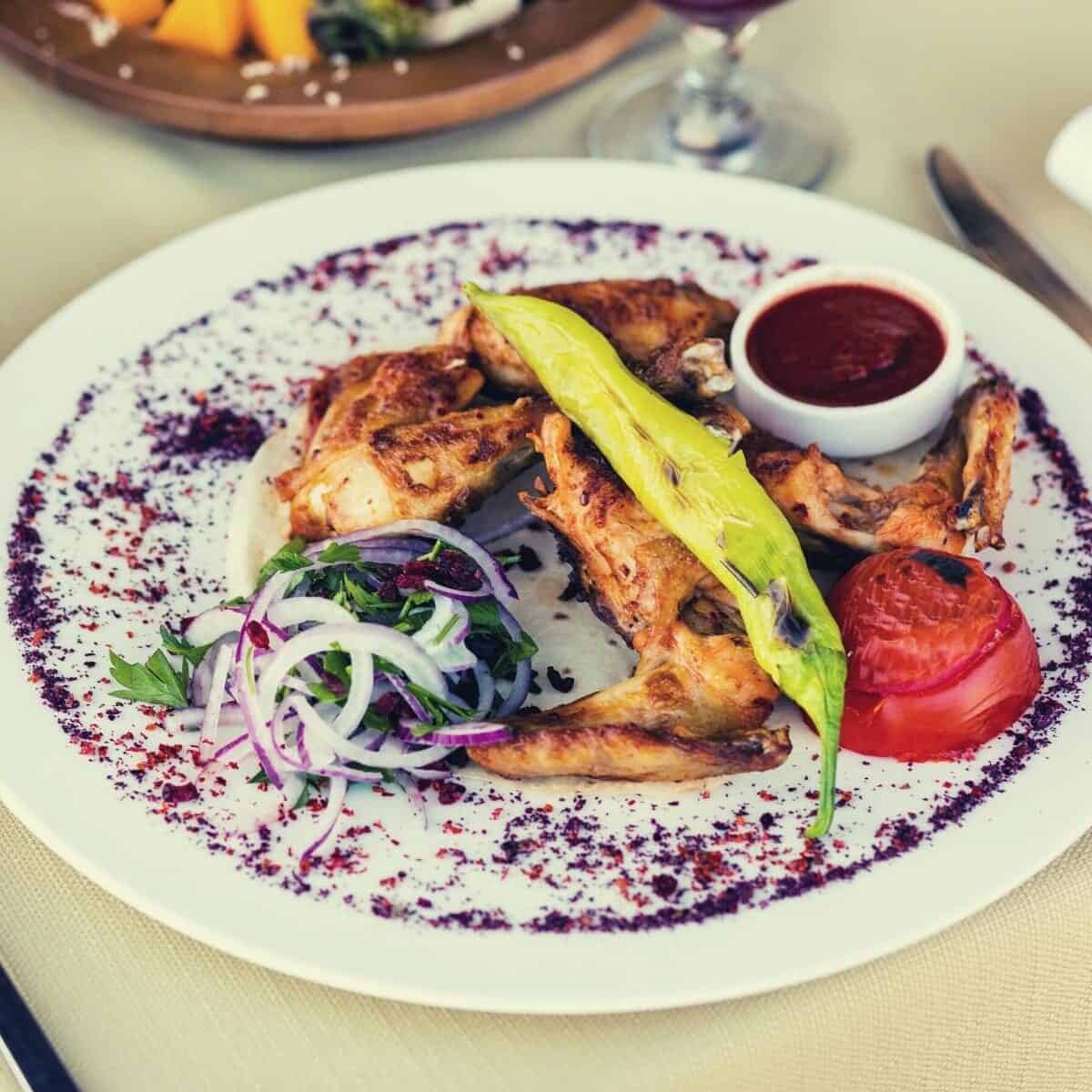 Grilled chicken wings, inspired by a classic Mississippi Chicken Recipe, are served on a white plate with a side of onion salad, grilled tomato, green chili, and a small dish of red sauce.