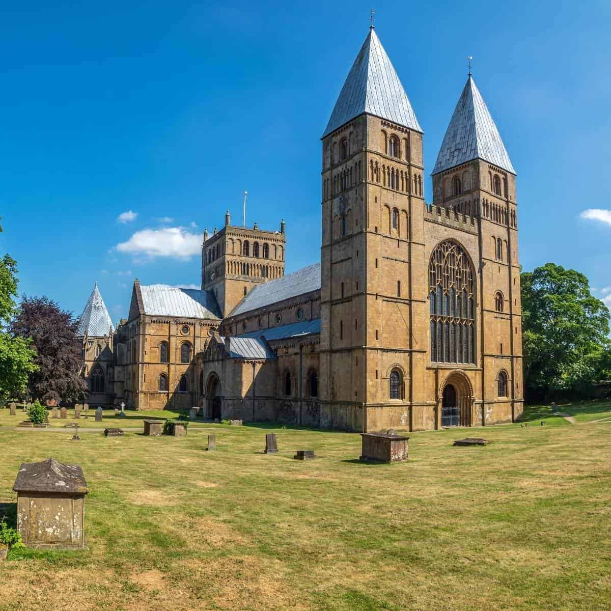 A historic stone cathedral in Nottinghamshire boasts two tall towers and large arched windows, set in a grassy yard with scattered gravestones under a clear blue sky—a must-see for those exploring on a budget.