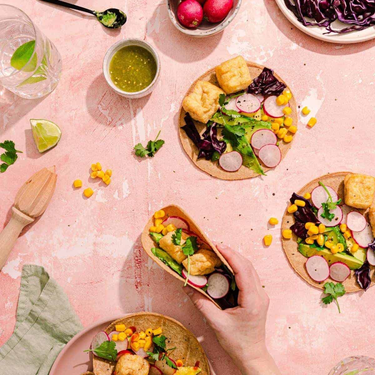A table showcases three vibrant vegan tacos filled with tofu, radishes, corn, and greens. A hand gently lifts one wrap. Nearby are fresh radishes, a lime wedge, green sauce, and a refreshing glass of water—perfect accompaniments to this delightful recipe.