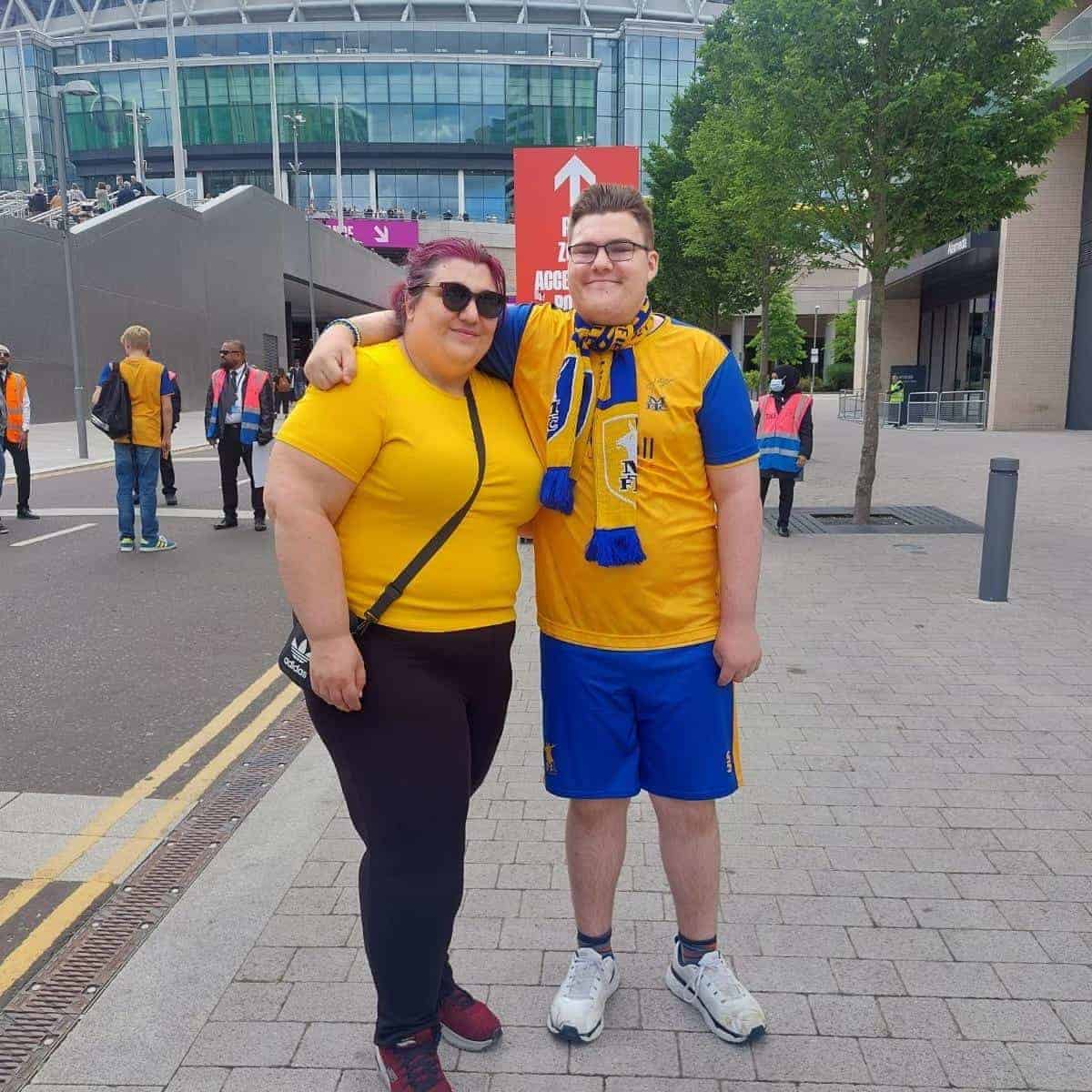 Two people in yellow shirts stand cheerfully together outside a stadium; one sports a scarf. Update from me: both are beaming as other fans and trees fill the background, capturing the vibrant atmosphere.