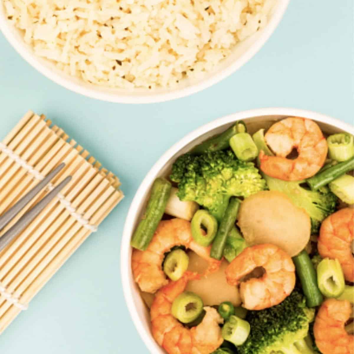 A bowl of sticky prawns stir-fry with broccoli and greens sits next to jasmine rice on a light blue surface, accompanied by chopsticks.